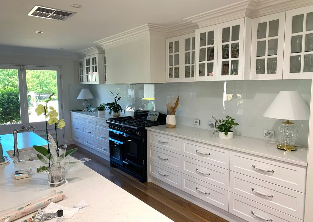 White Hamptons-style kitchen with glass-front cabinetry, black range cooker and glass splashback